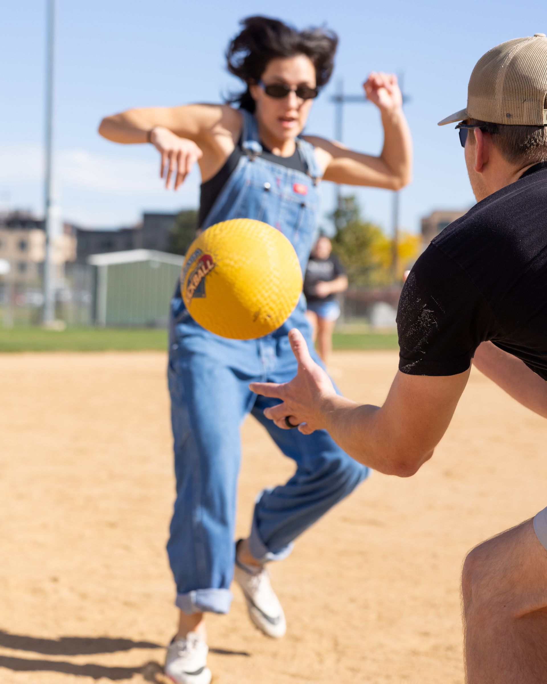 Woman in overalls kicks yellow ball; another person attempts to catch it on a sunny field.