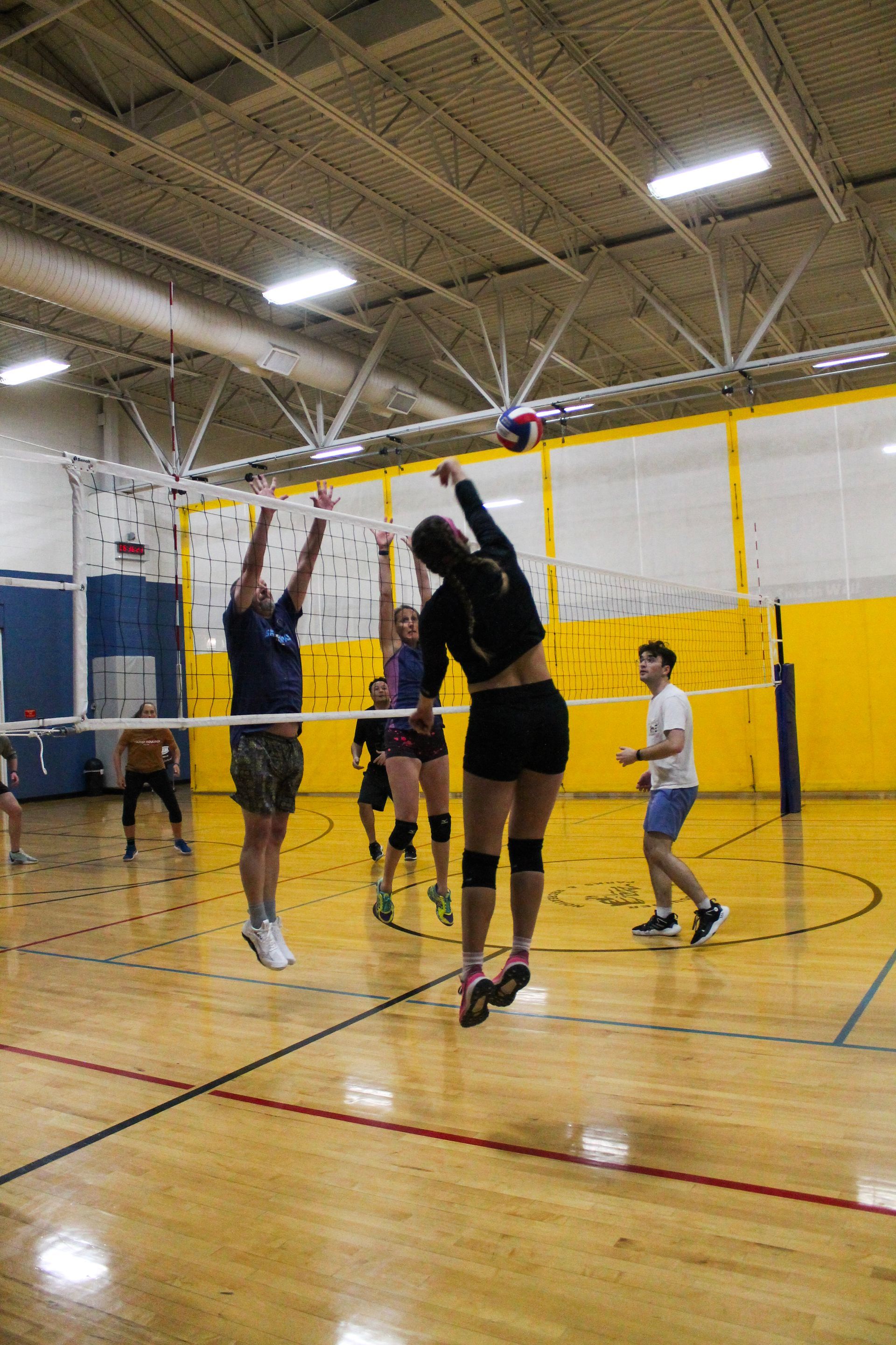 Volleyball game in a gym. Player spikes the ball over the net while others jump to block. Yellow and blue walls.