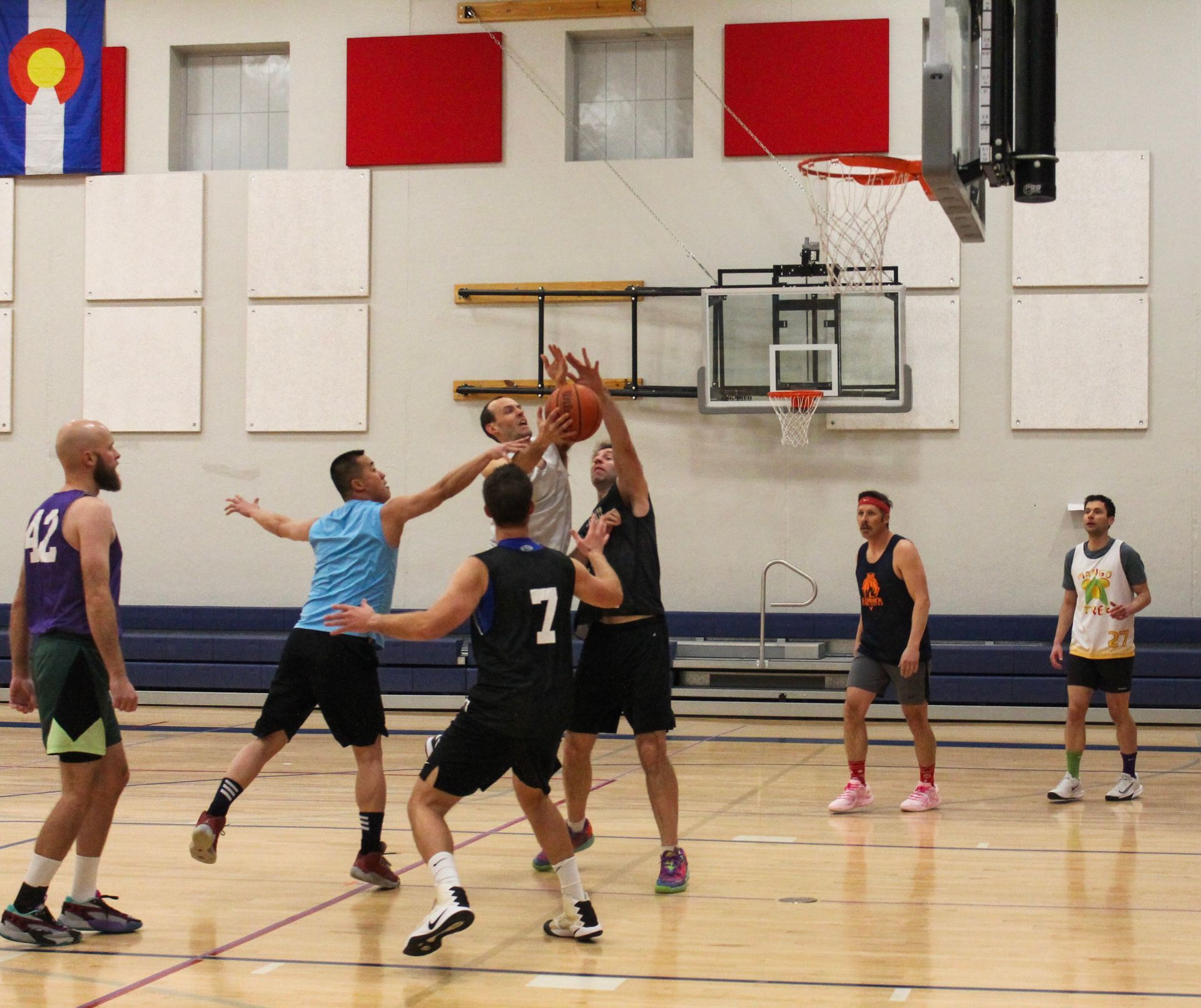 Basketball game in a gym. Players in jerseys compete for a rebound. Colorado flag in the background.