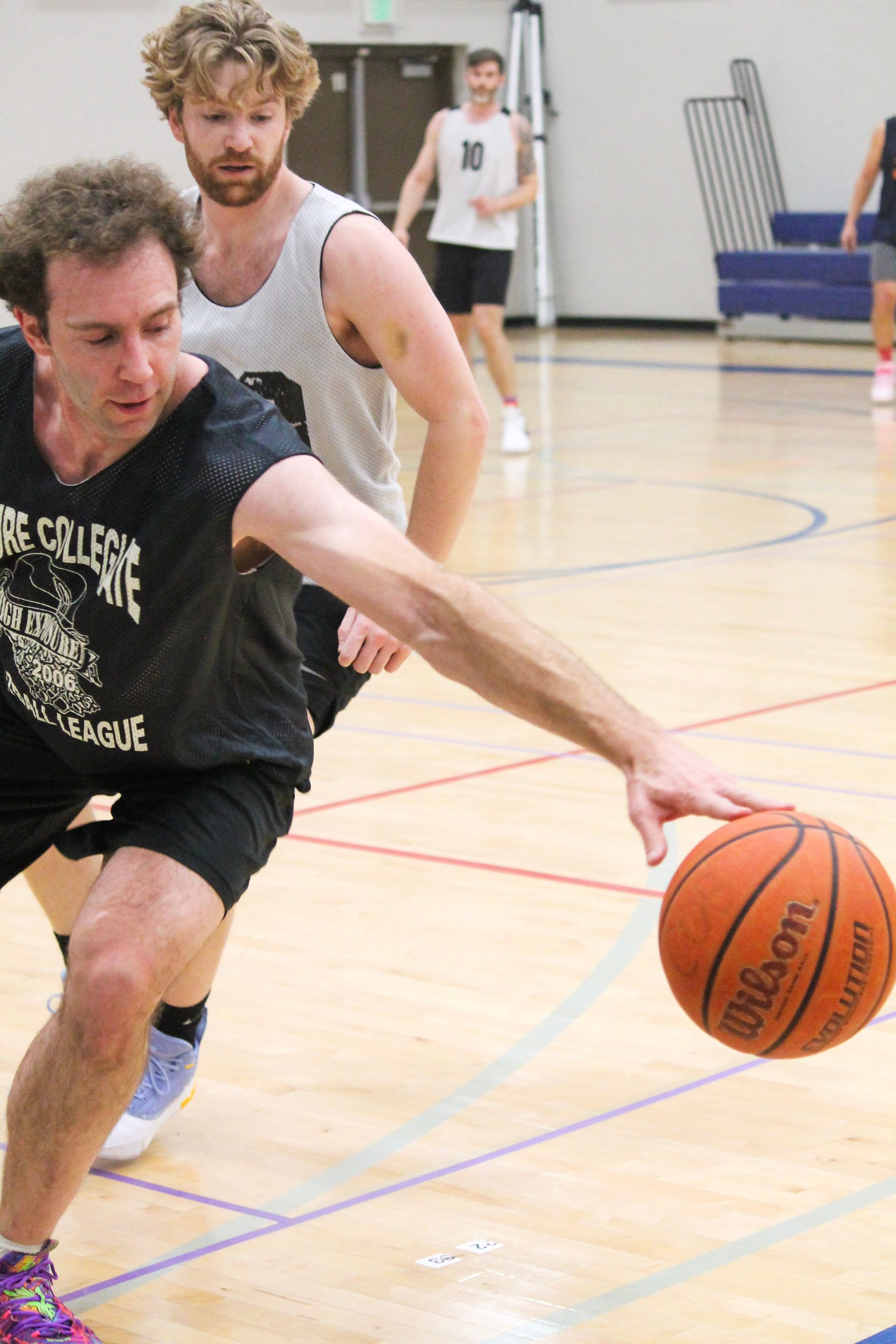 Man dribbling a basketball on a court, guarded by another player. Other players in the background.