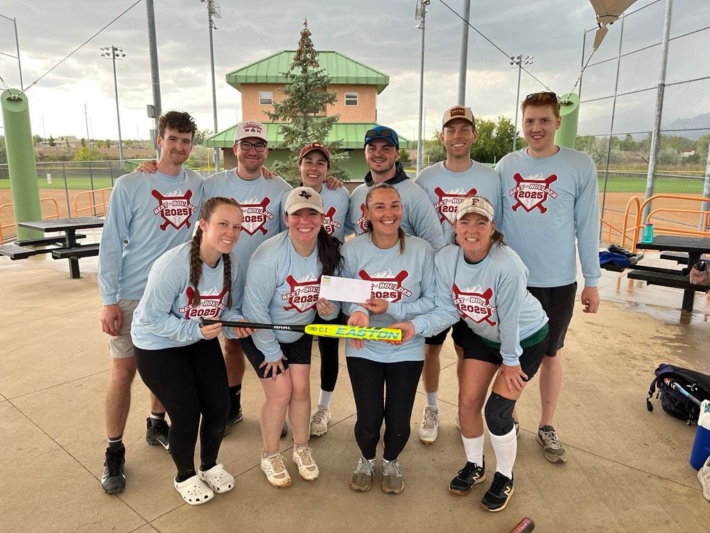 Softball team in matching light blue shirts holding a large check, posing for a photo at an outdoor park.