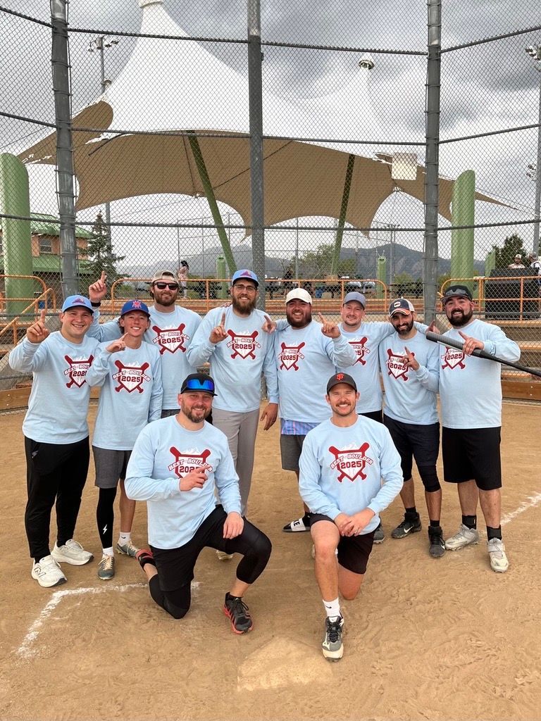 Softball team in light blue shirts poses on a dirt field with a netting enclosure.