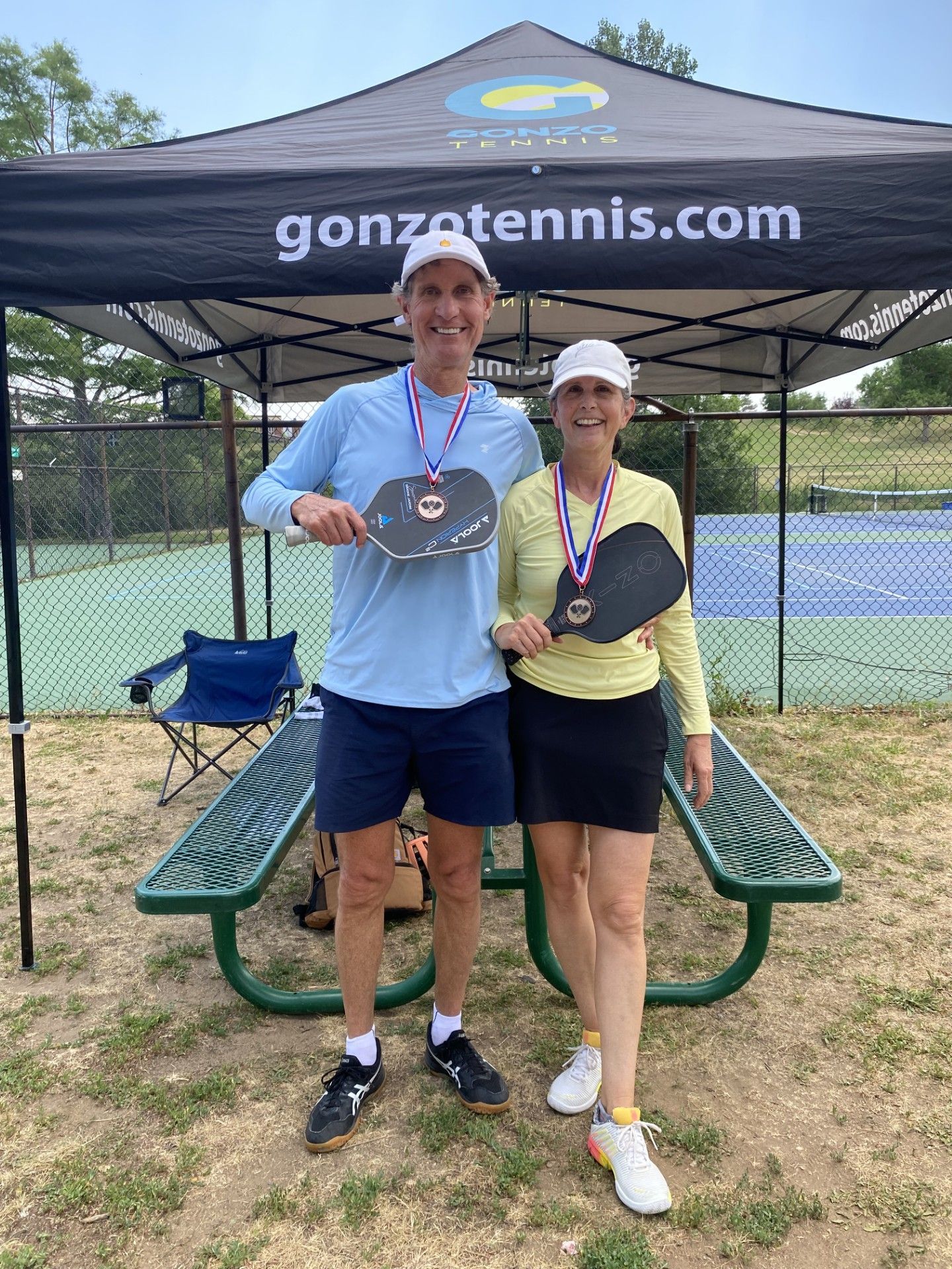 Man and woman holding pickleball paddles, wearing medals, under a tent at a tennis court.