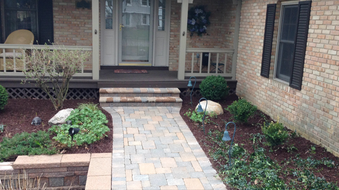 Brick pathway leading to a house with landscaping and a dark brown door and trim.