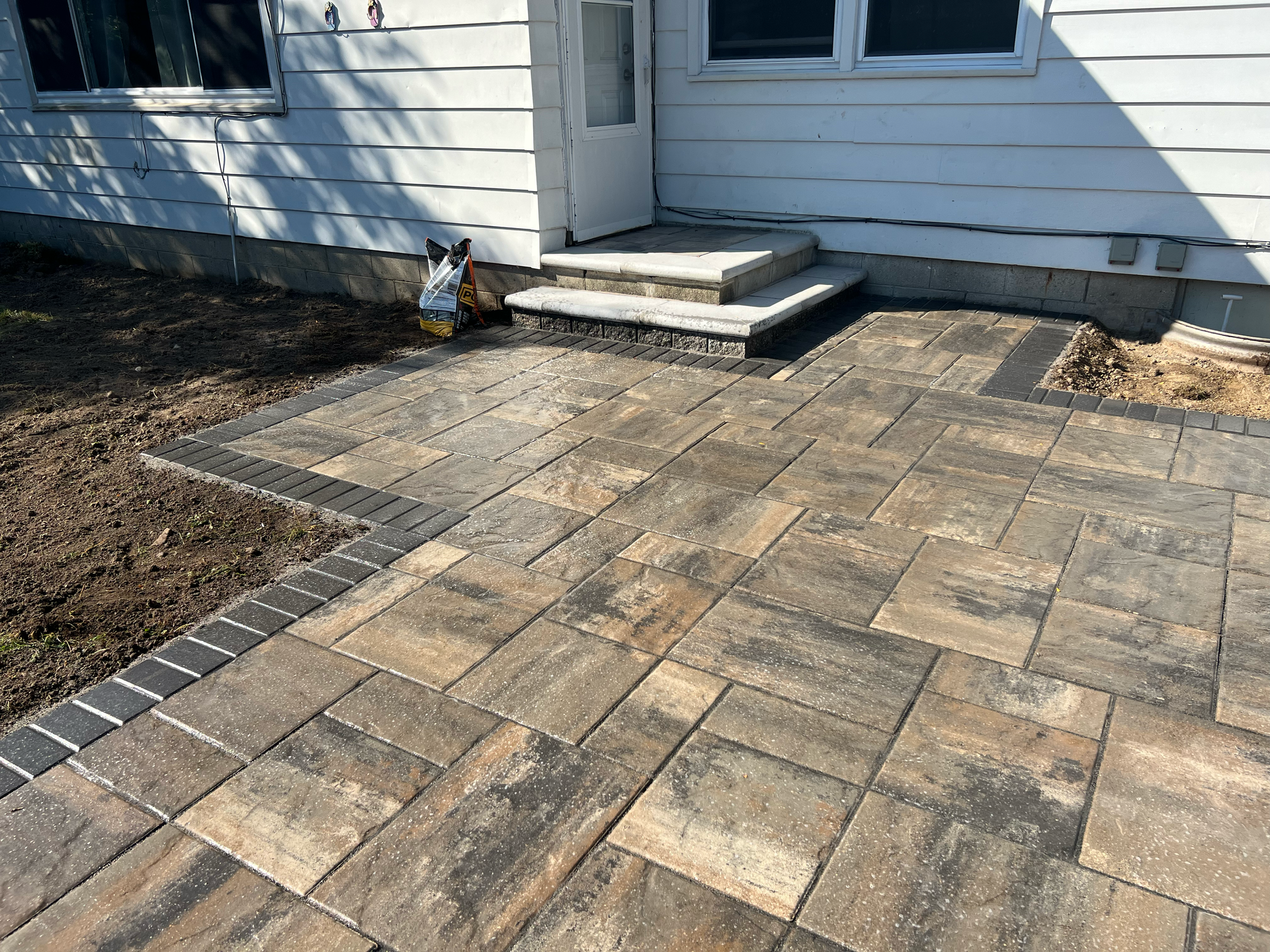 Brick patio with steps leading to a white house entrance. Bordered by a black brick edge.