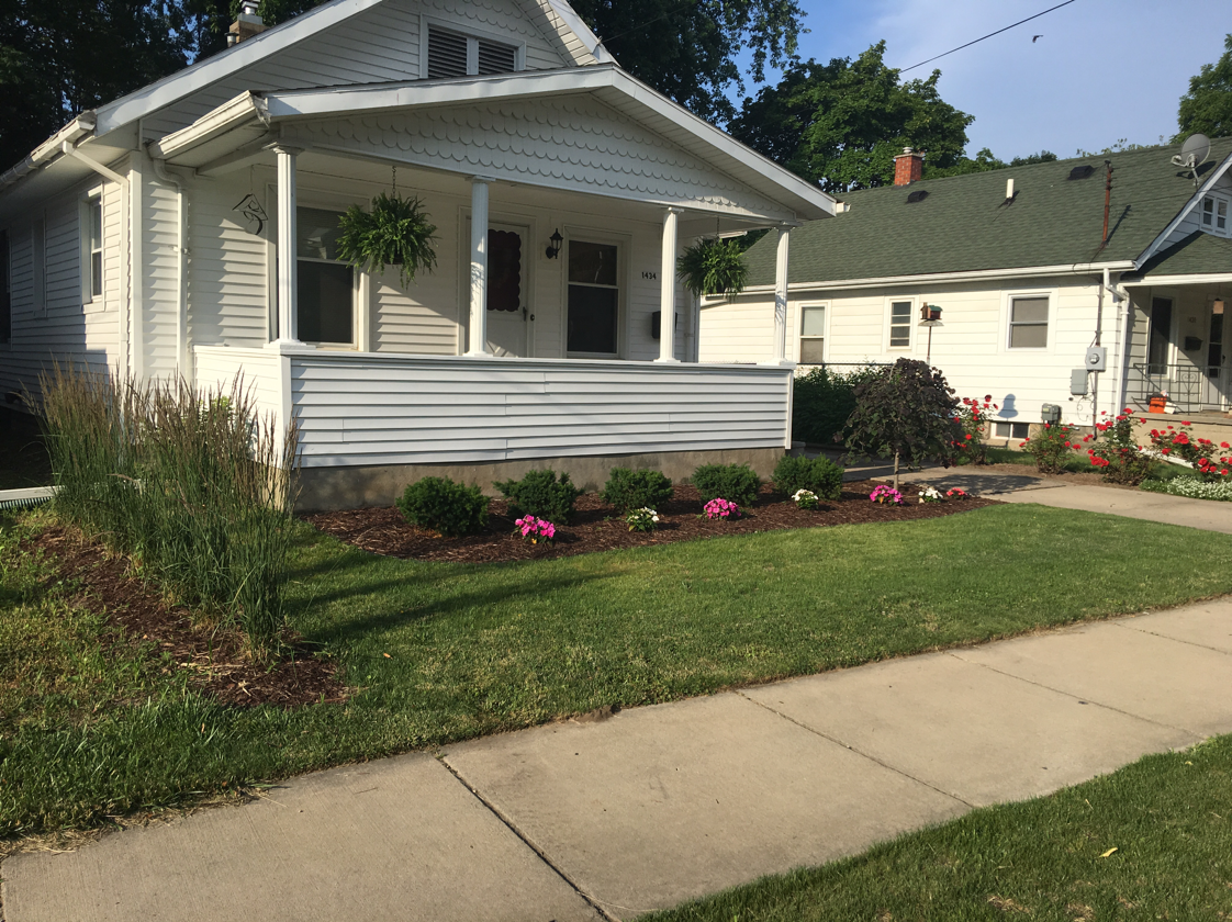 White house with porch, flower beds, and green lawn next to a sidewalk.
