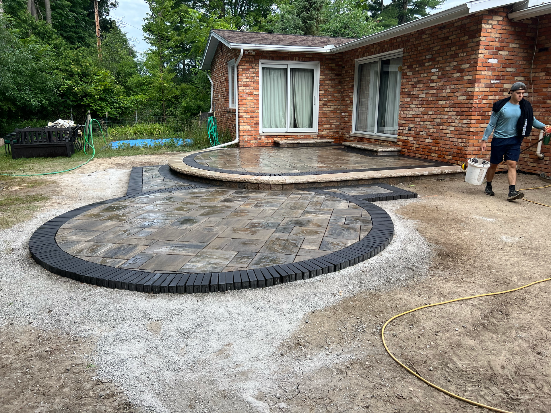 Brick house with newly constructed brick patio and person carrying bucket. Gravel and trees in background.