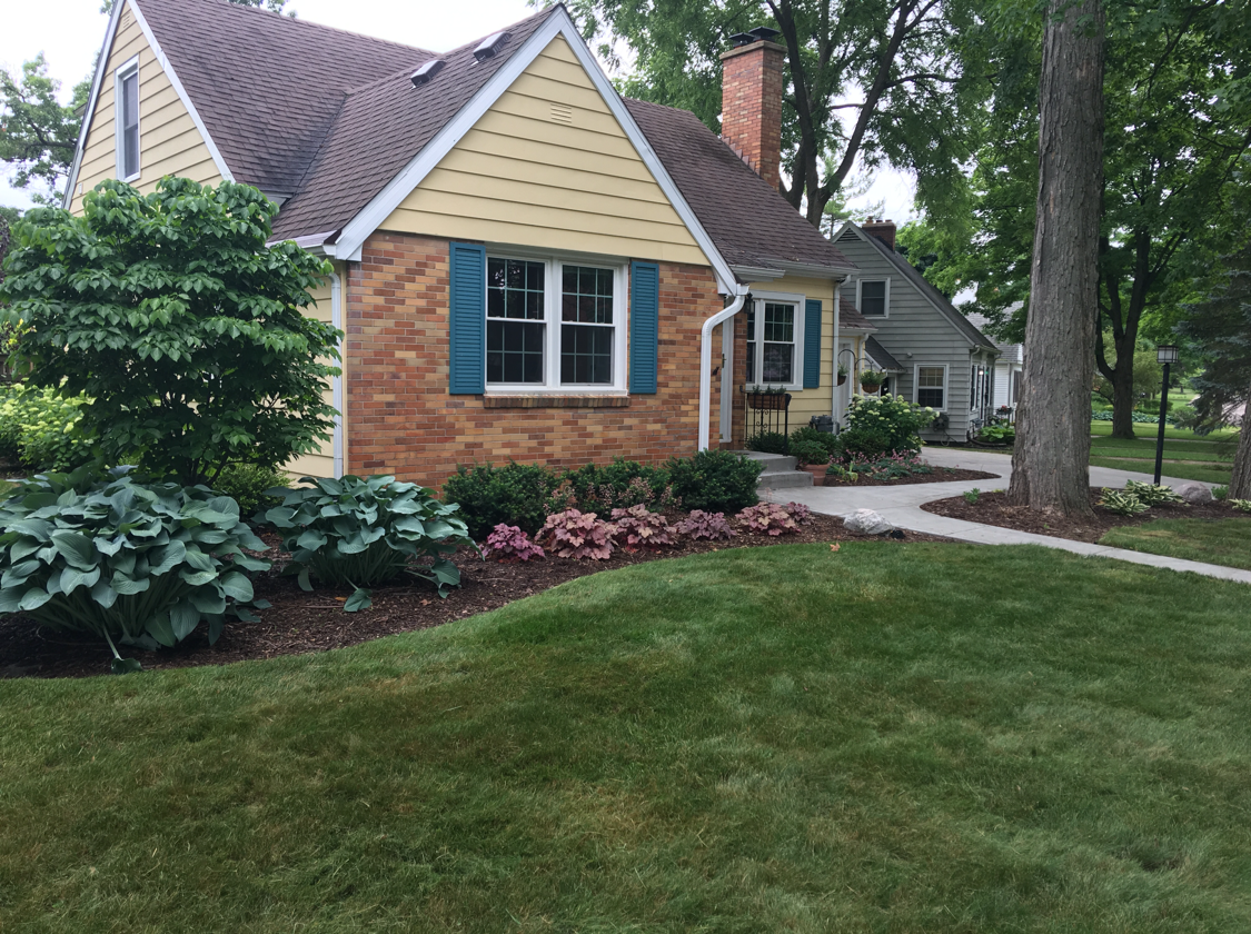 Yellow house with blue shutters, brick, and a well-maintained lawn and garden.