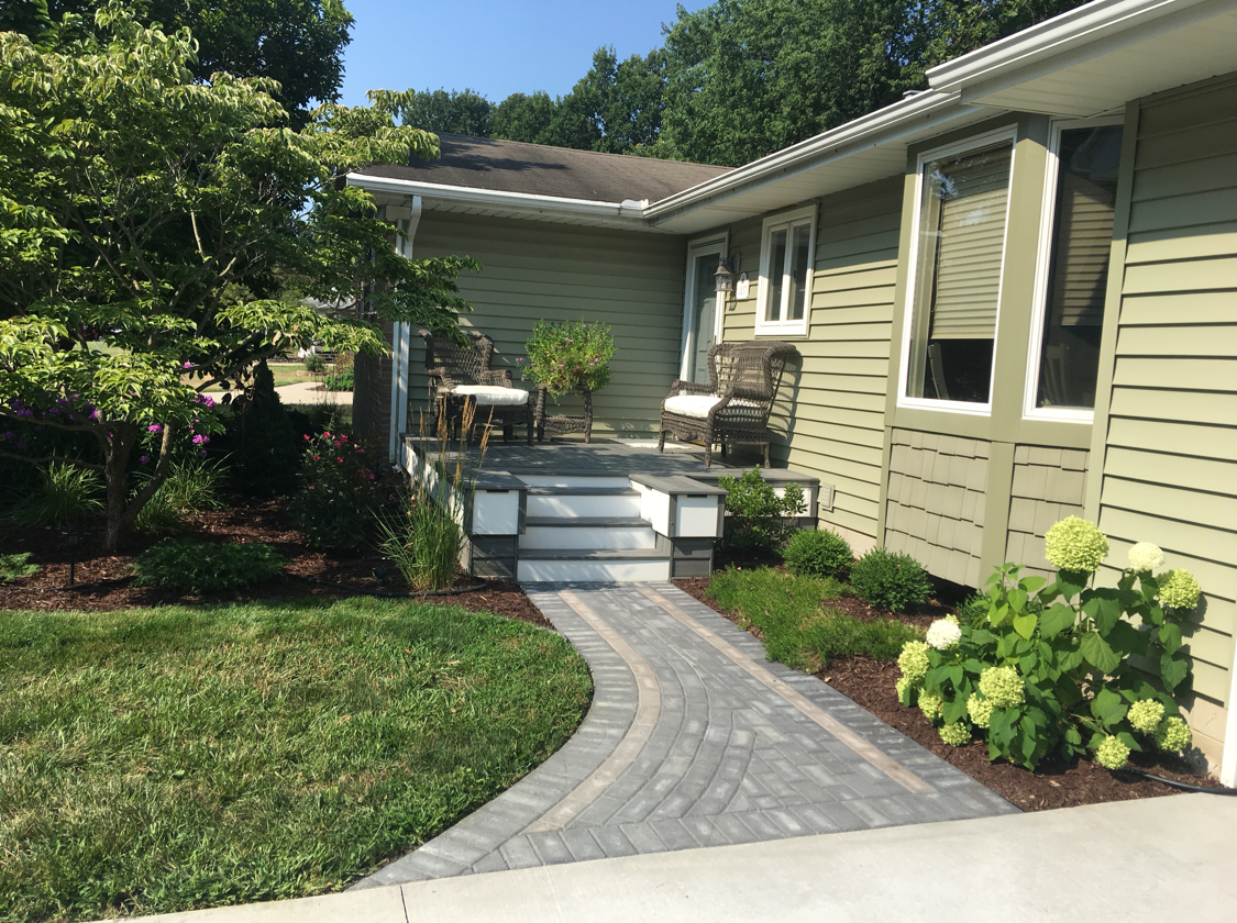 Stone pathway leads to a porch with chairs. Green siding, landscaping, and blue sky.