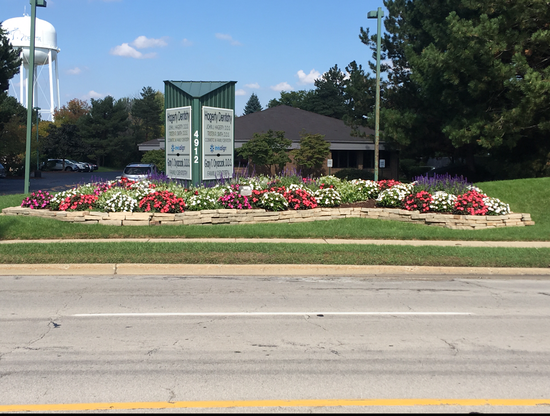 Landscaped sign with colorful flowers, water tower, and building in the background, along a road.