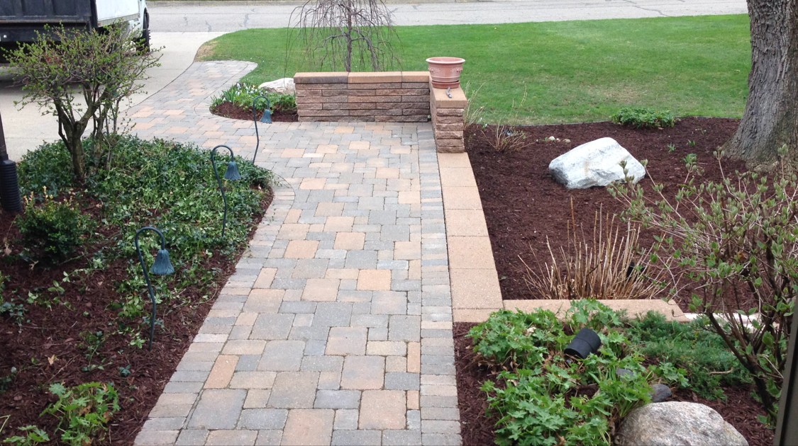 Brick walkway leading to a brick and stone wall. Landscaping with mulch, plants, and a large rock.