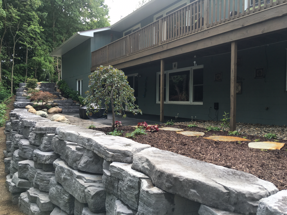 Stone retaining wall in front of a house with a deck; a small tree and landscaping is in between.