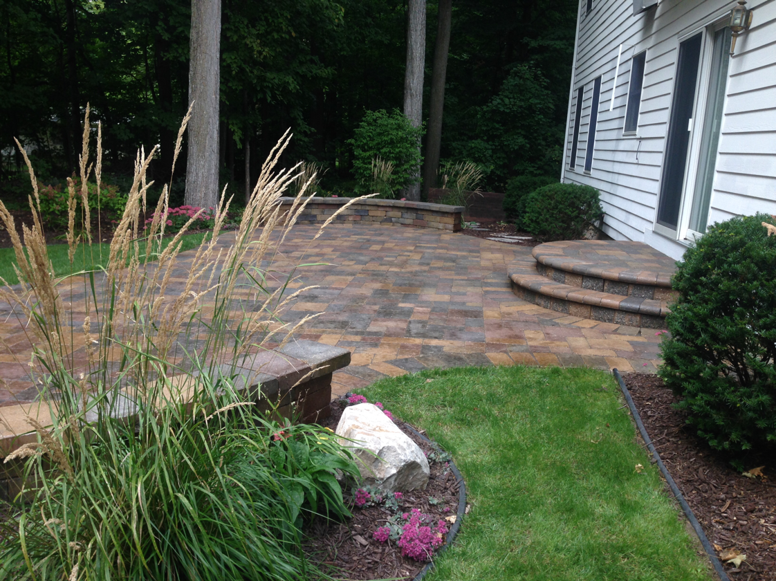 Brick patio with steps next to a white house, surrounded by grass and landscaping.