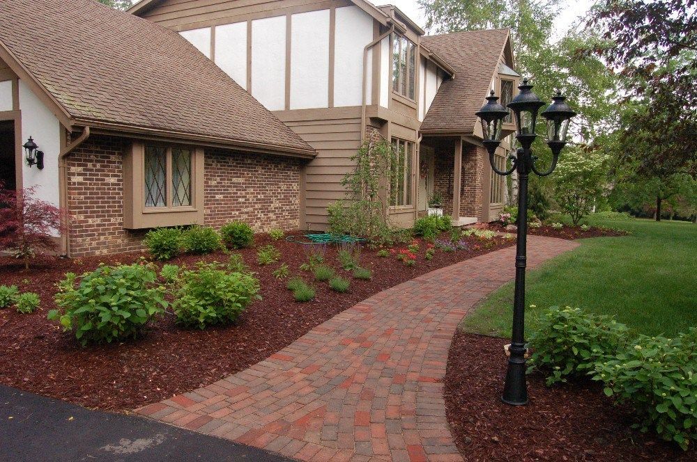Brick pathway leads to a house with brown roof and trim, black lamppost, flowerbeds with reddish mulch.