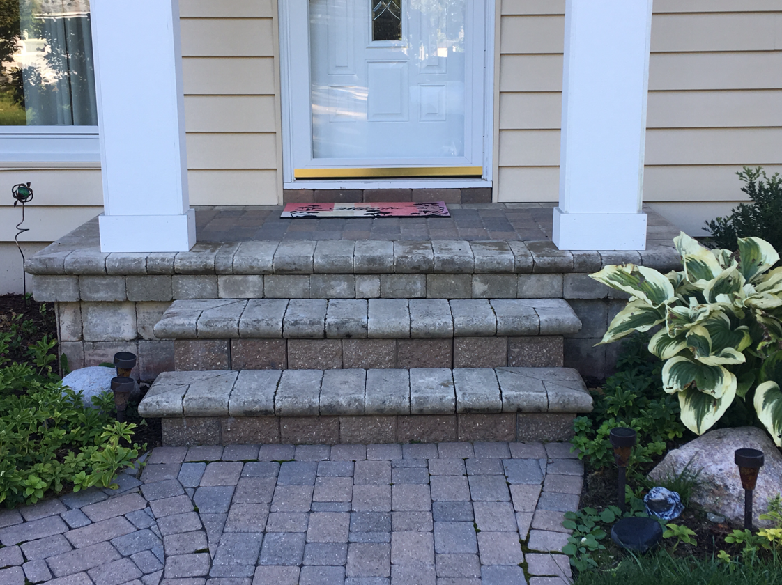 Brick steps leading up to a beige-sided house with white columns and a front door. A brick path leads to the steps.