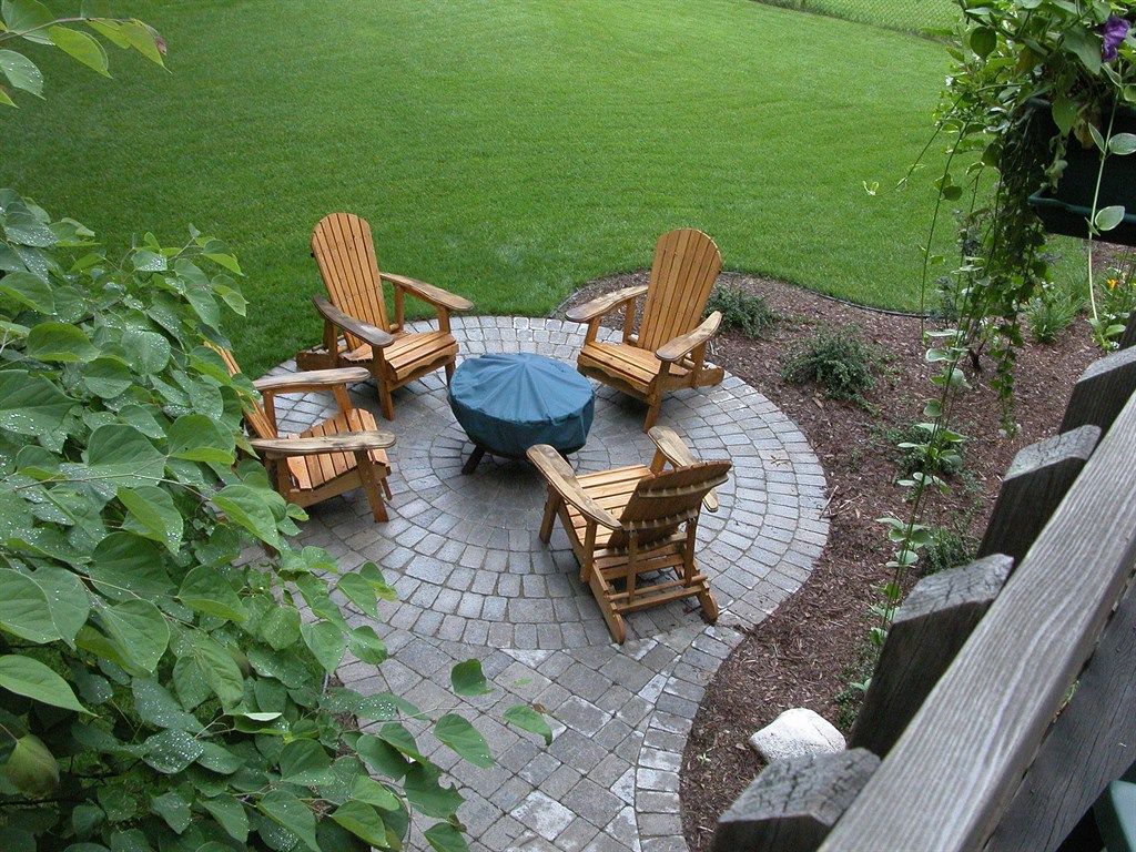 Four wooden chairs surround a covered fire pit on a brick patio, set in a green lawn.