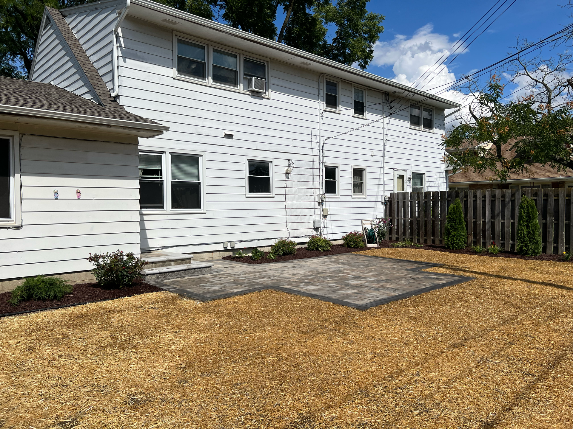 Backyard with white house, paved patio, brown mulch, and wooden fence.
