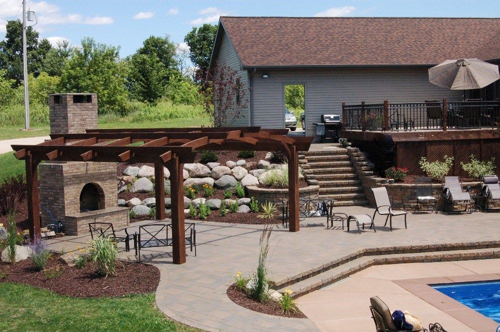 Outdoor patio with pergola, fireplace, pool, and deck. A house with a brown roof sits in the background.