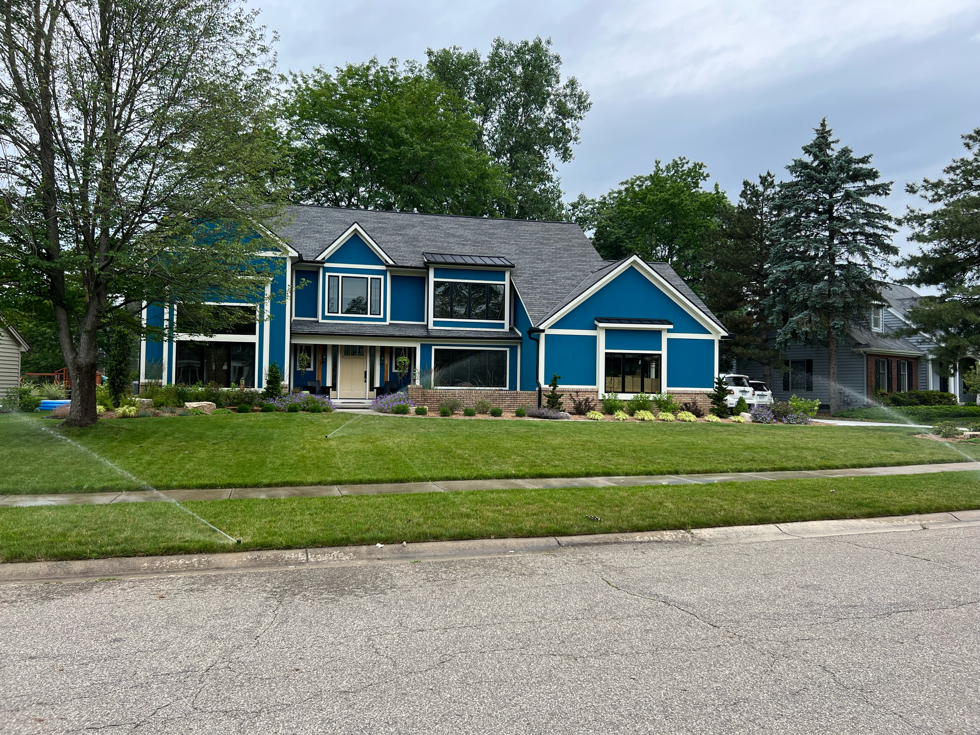 Blue house with dark gray roof and manicured lawn. Sprinkler system spraying water.