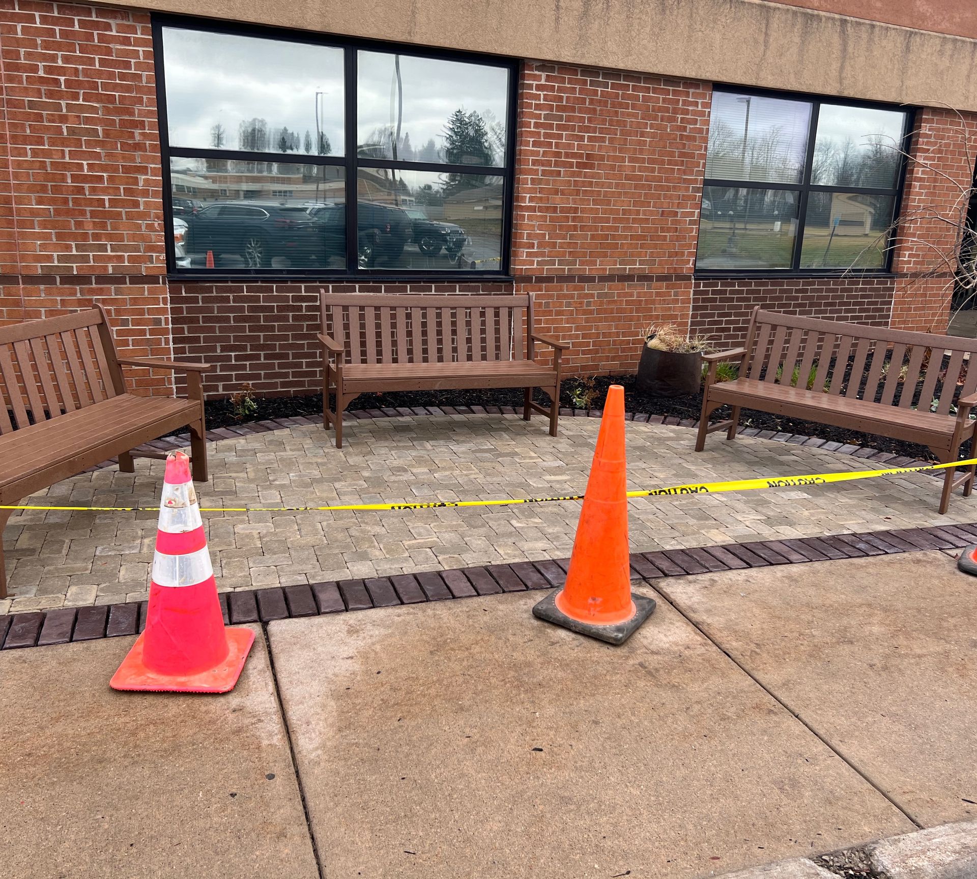 Three wooden benches, orange cones, and yellow caution tape on a brick and concrete surface outside a building.