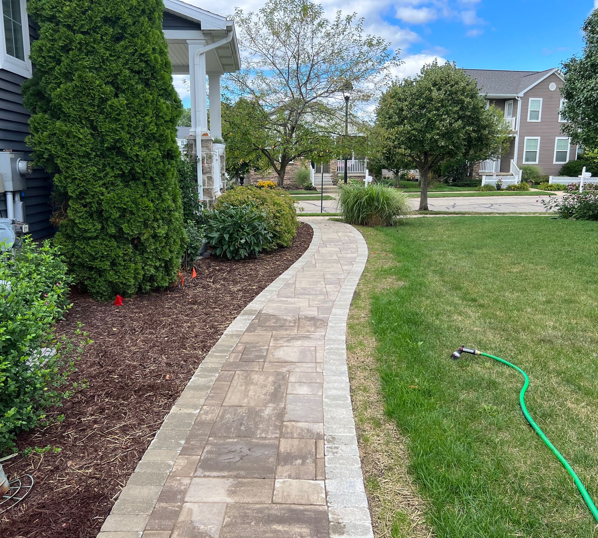 Brick pathway curves through a yard, leading toward a house with dark siding; green grass on the right.