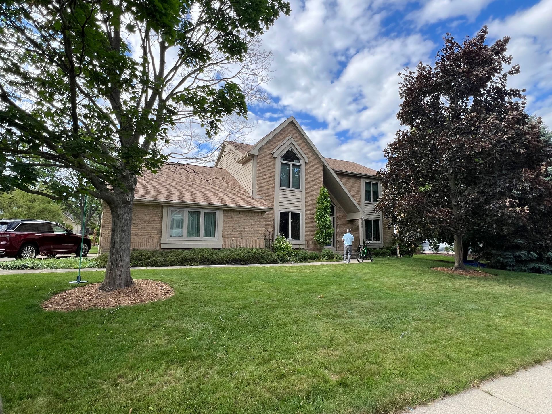 House with brick exterior, green lawn, trees, and a person standing in front. Overcast sky.