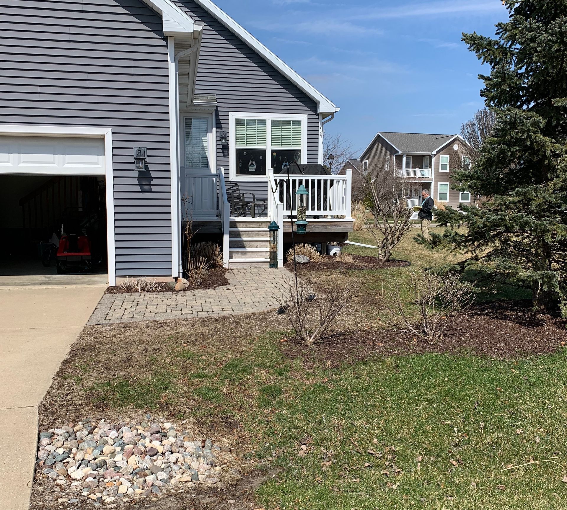 A house exterior with a gray garage, steps to a deck, and landscaping; sunny day.