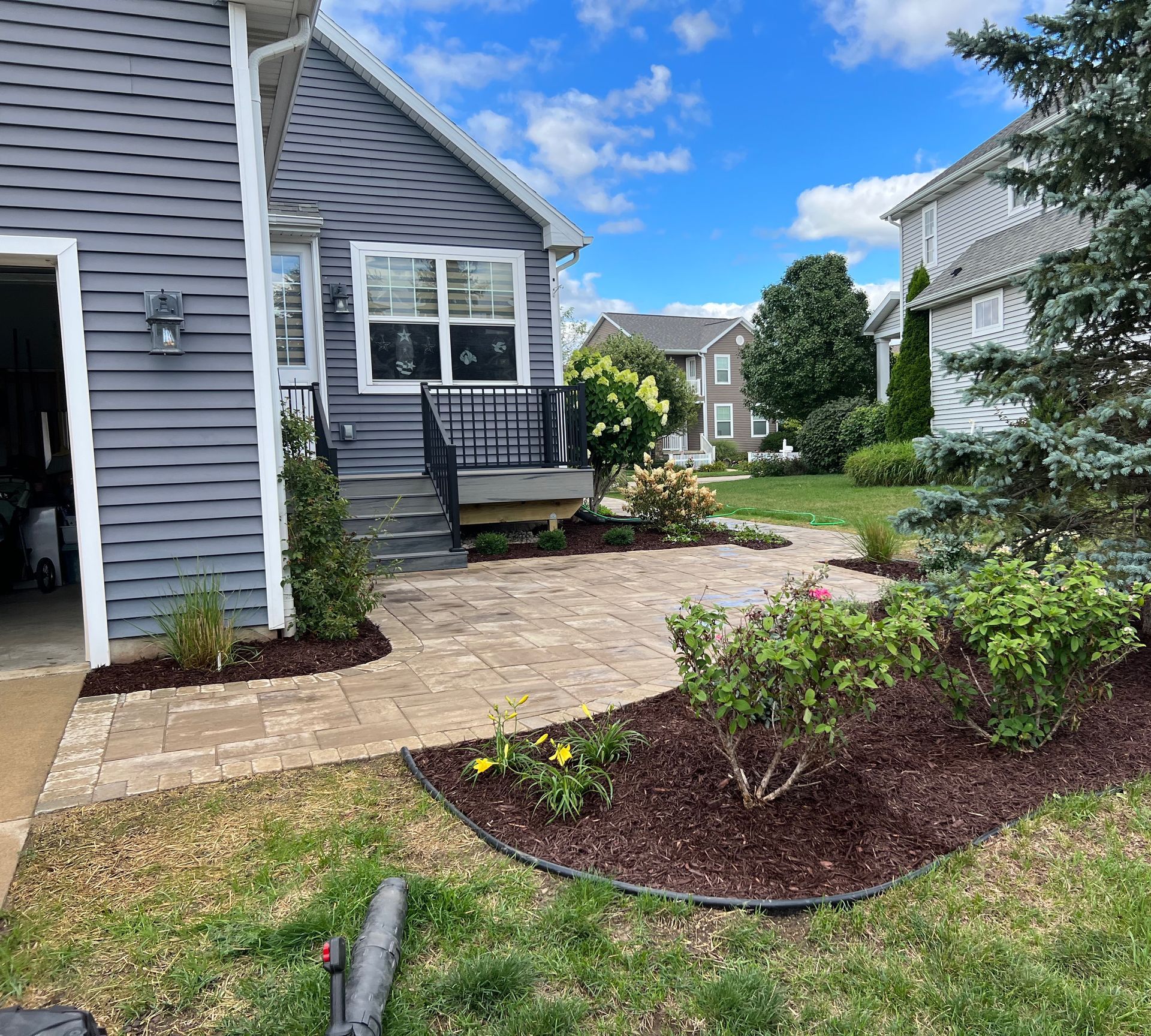 Brick patio with landscaping by a gray house and garage. Sunny day.