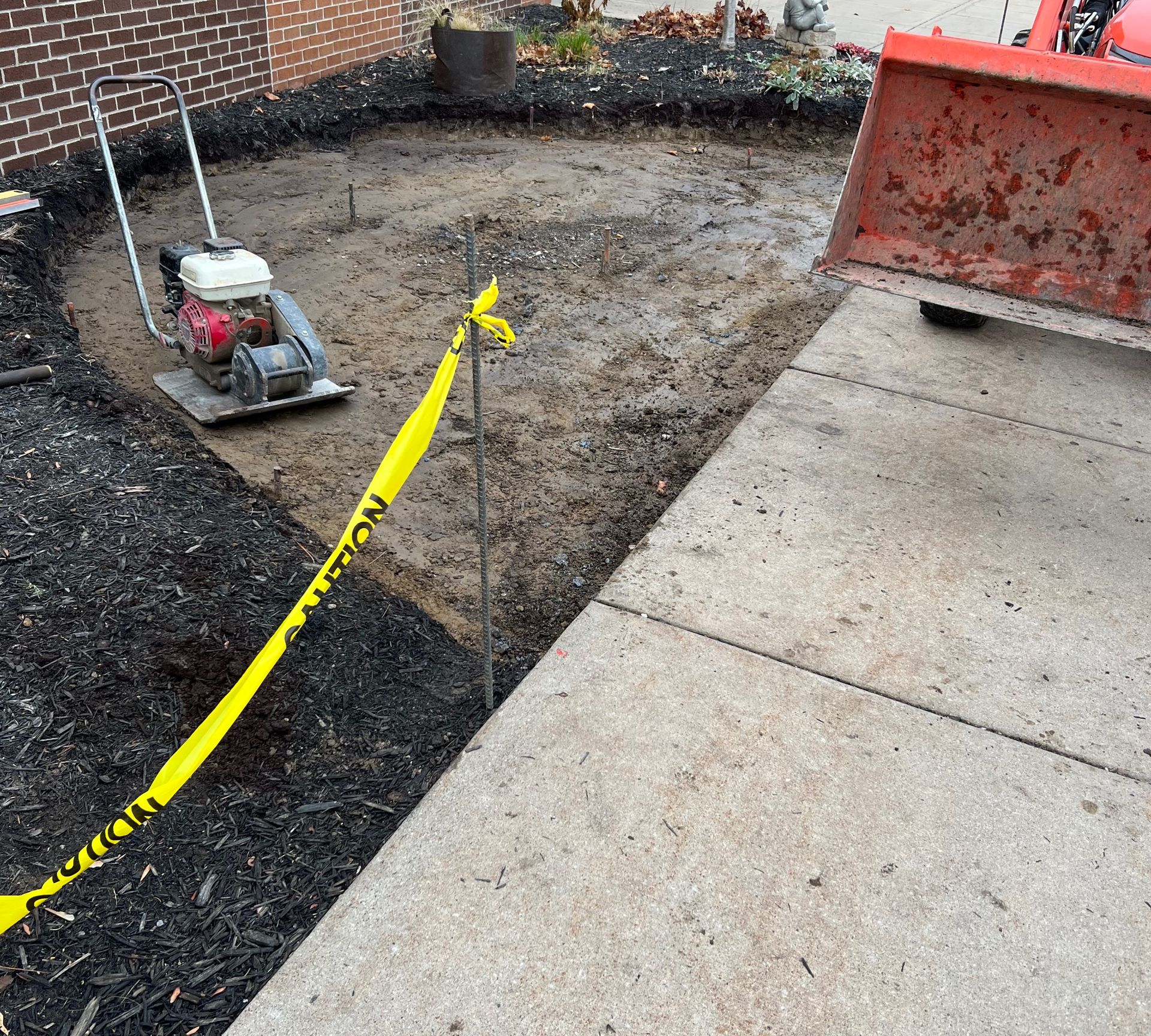 Compaction equipment and small tractor on a dirt area next to a concrete driveway, bordered by black mulch.