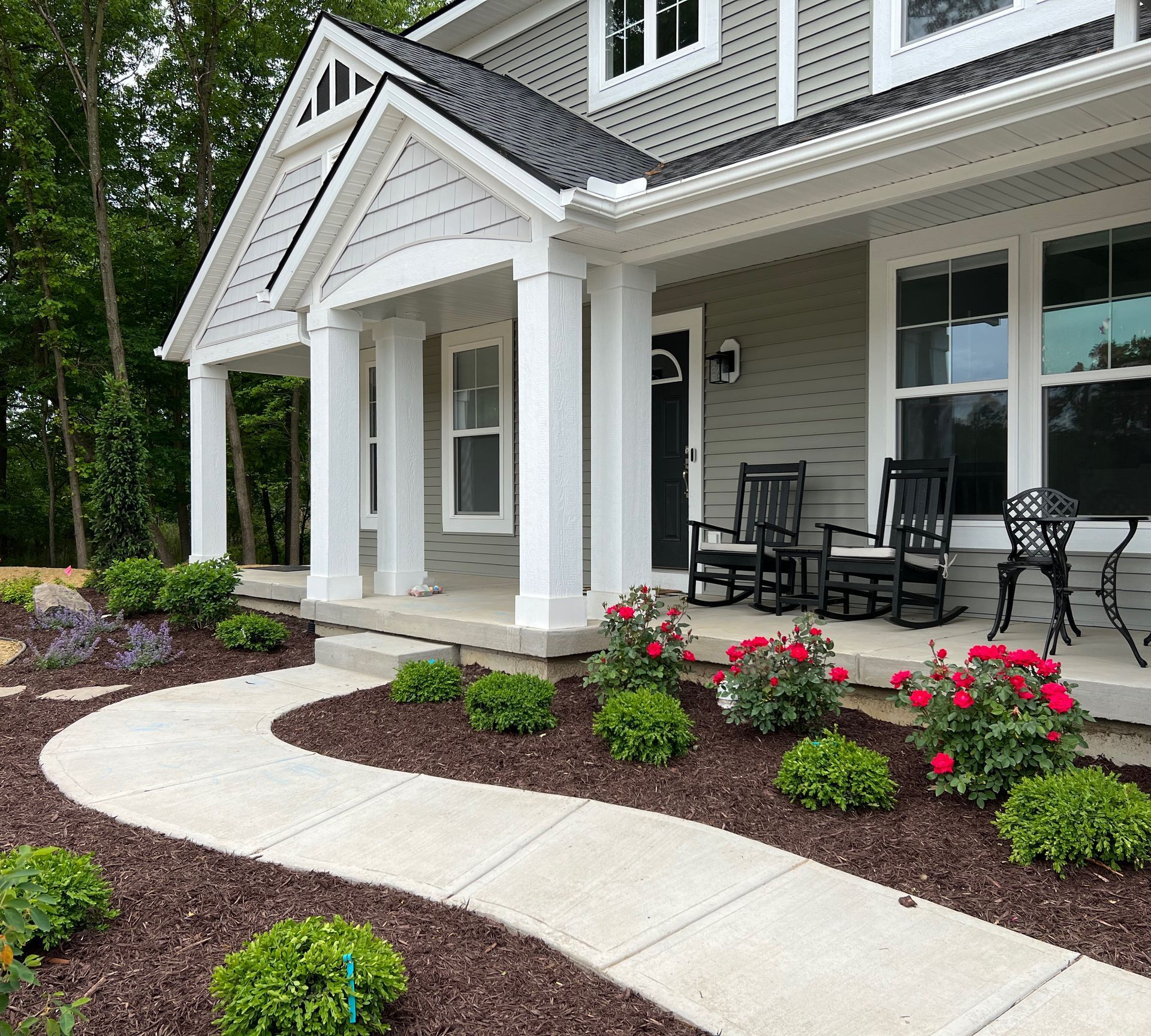 Front porch with black rockers, a winding walkway lined with flowers, and a gray house.