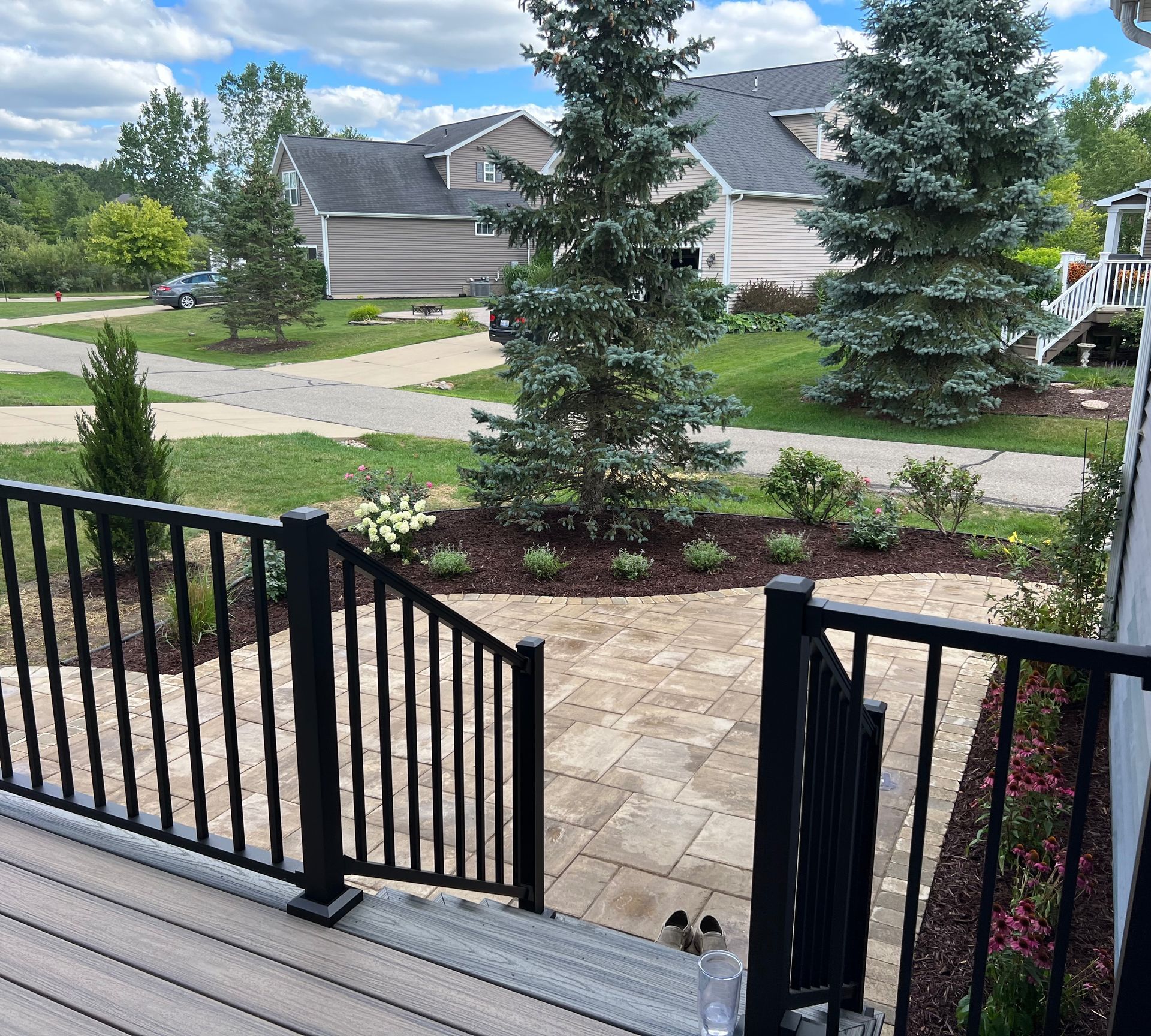 Black railing leads to a stone patio with landscaped flowerbeds and tall trees. Suburban setting.