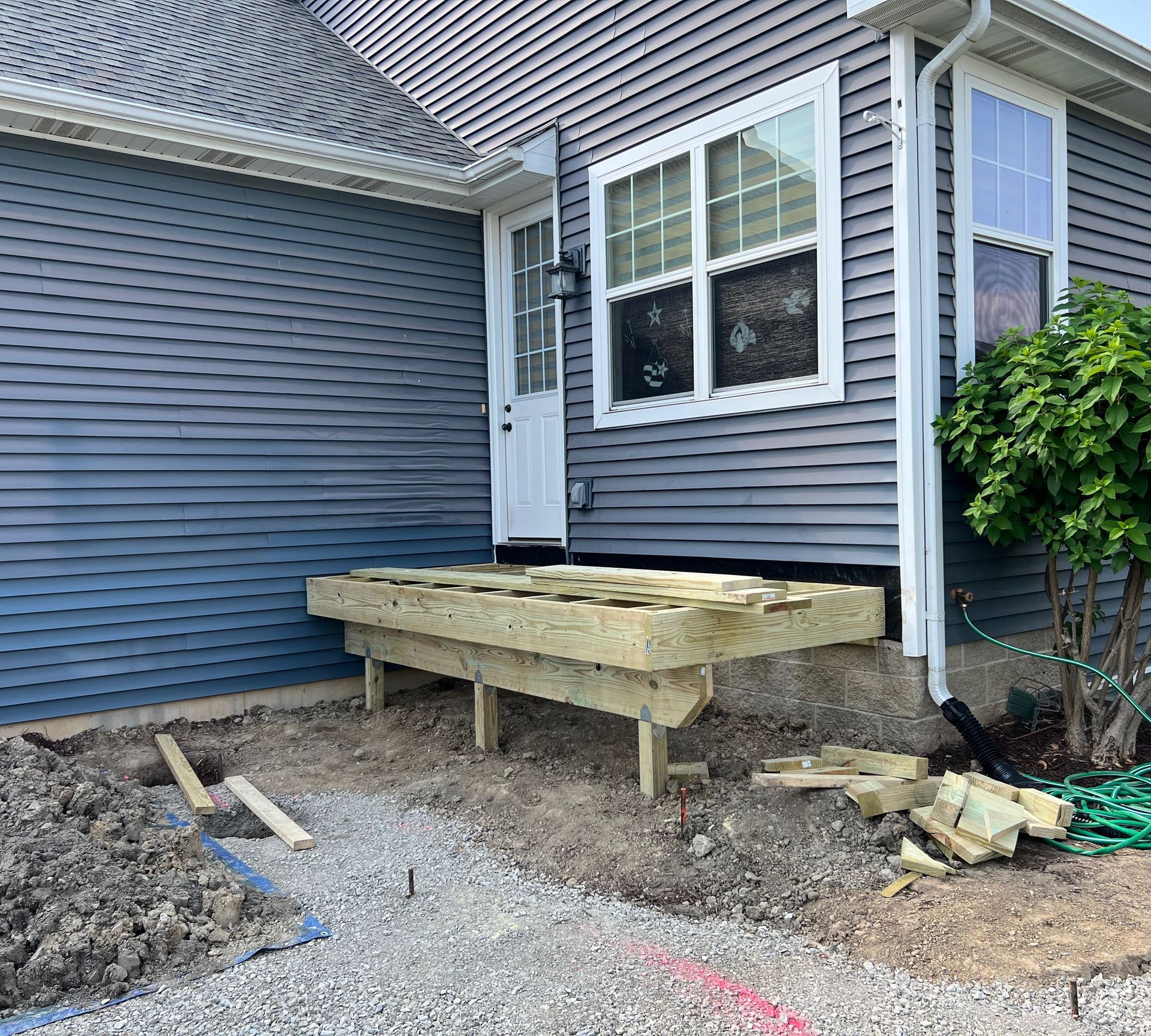 New wooden deck under construction outside a blue-sided house with a white door and window. Gravel and dirt are visible.
