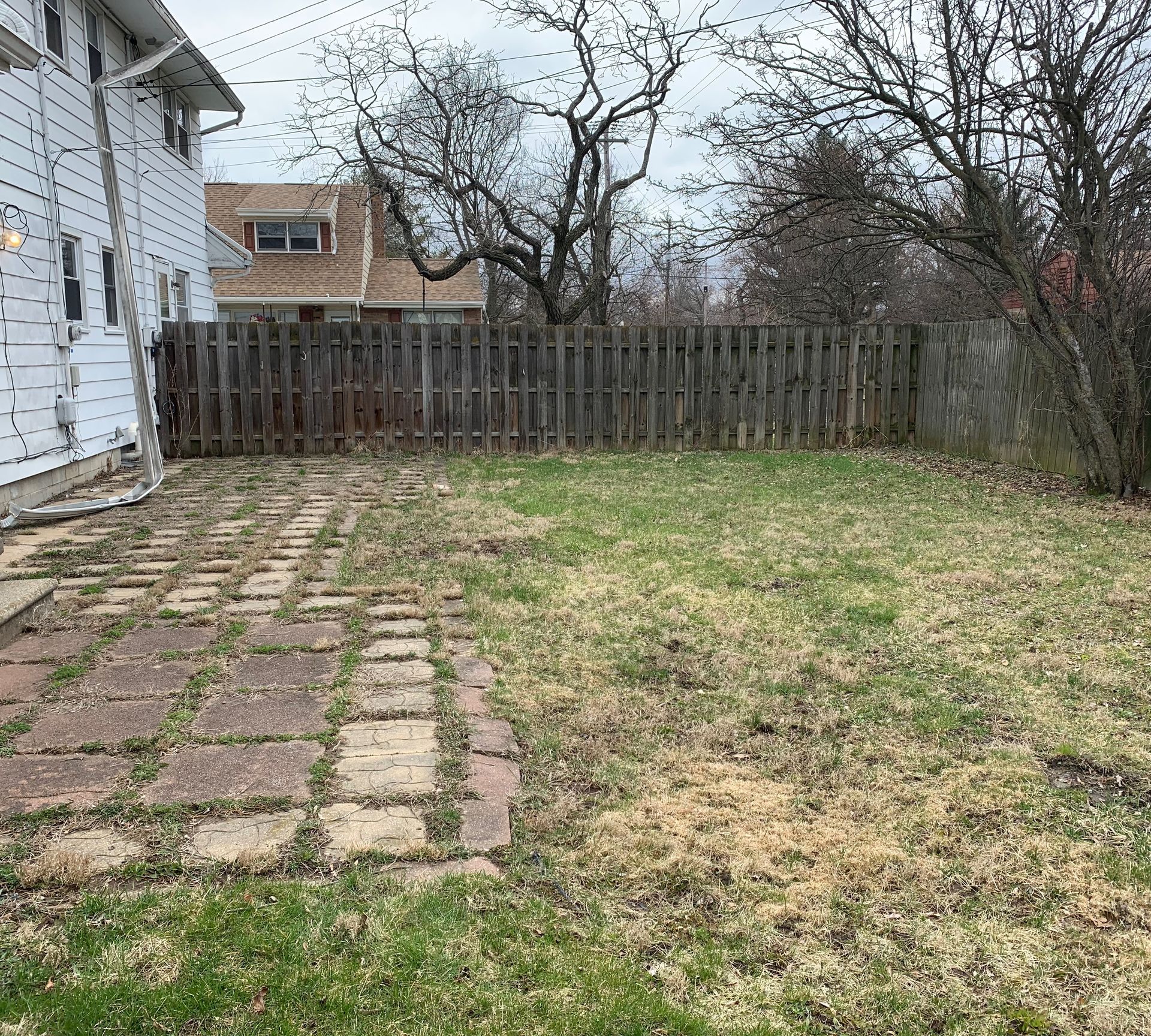 Backyard with a brick patio and fenced lawn, a house on the left, and bare trees in the background.