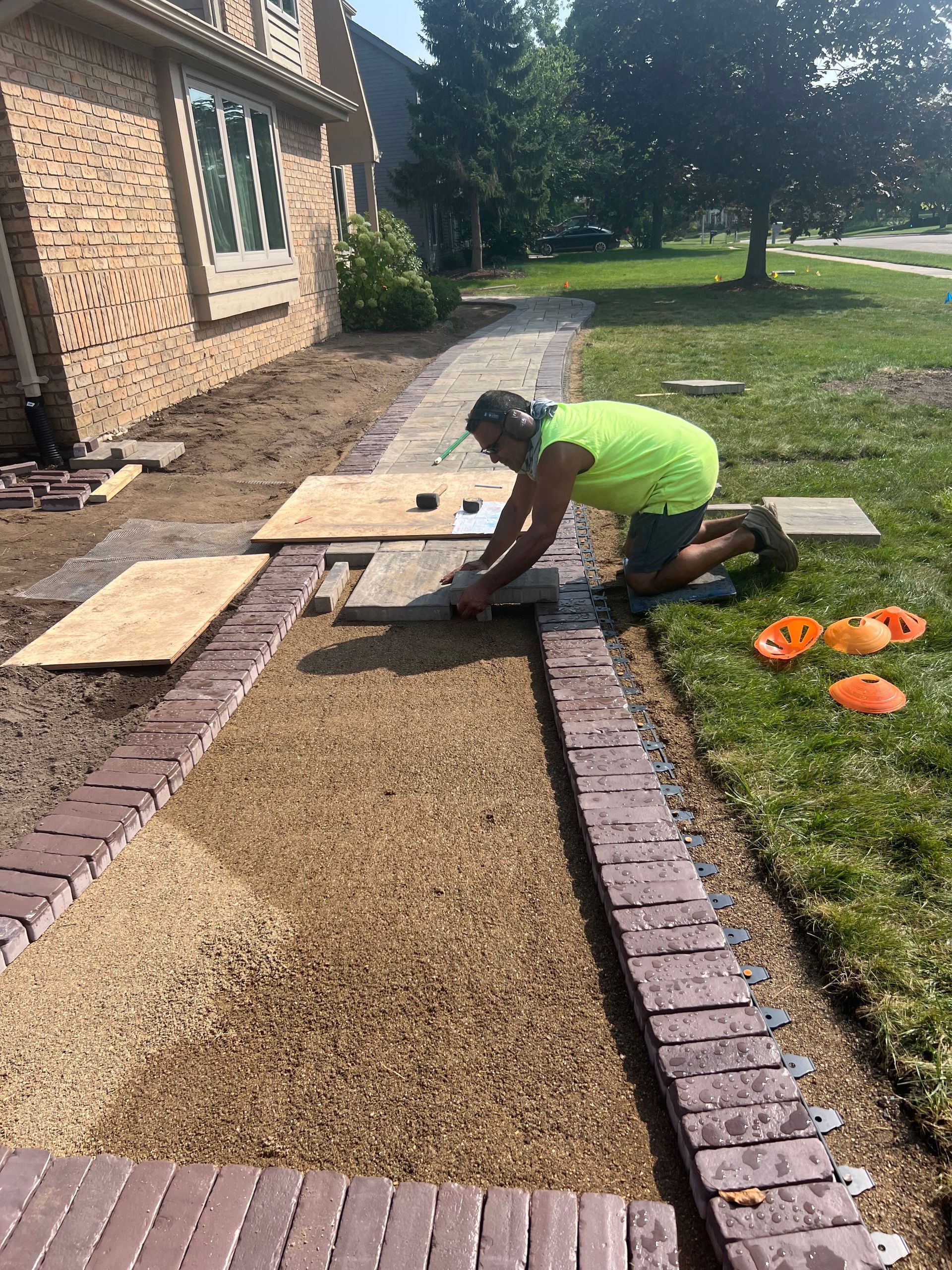 Man laying a brick walkway, kneeling on grass. Light brown gravel, red bricks, house in background.