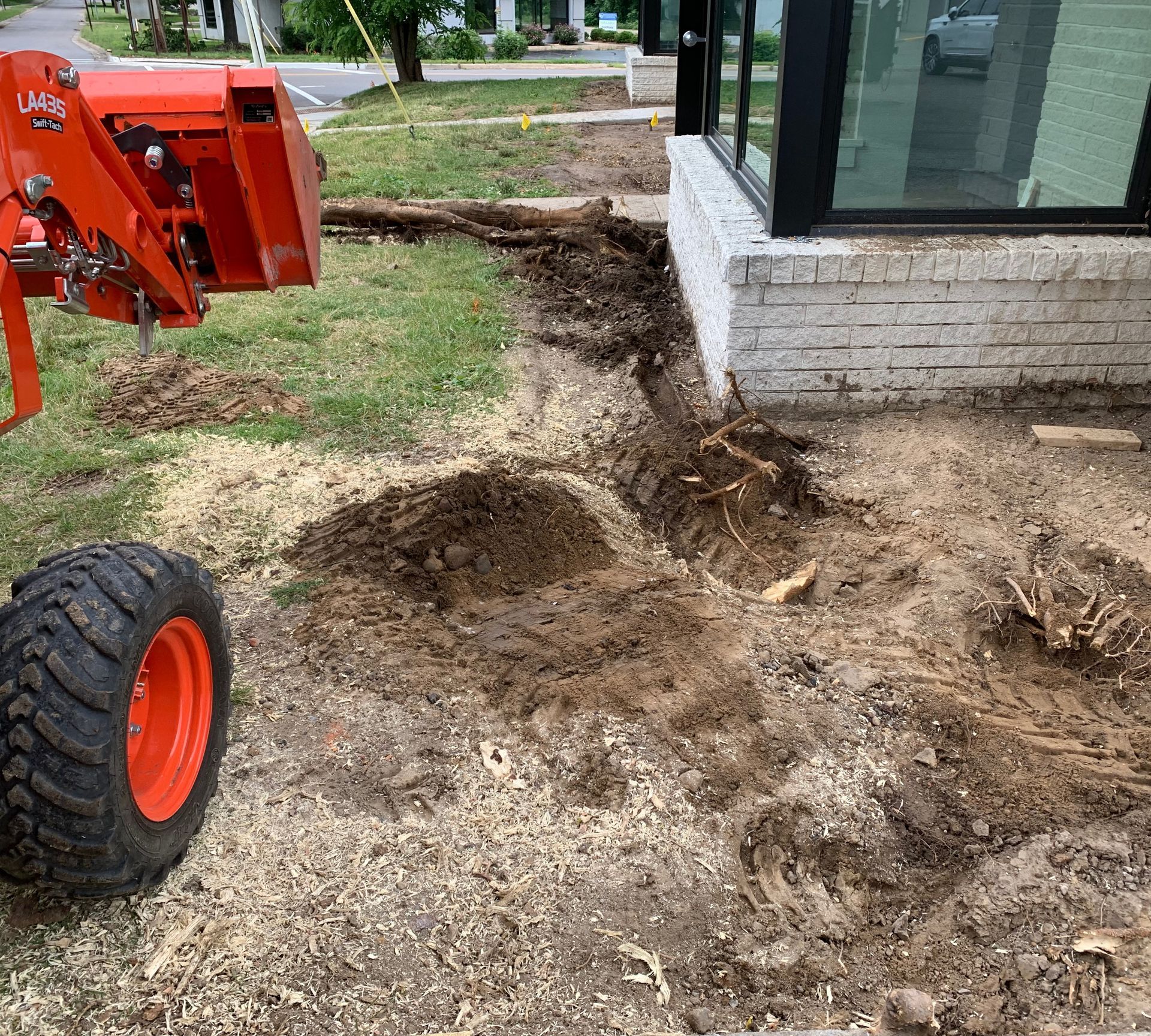 Orange tractor digging in dirt near a building with a glass window; outdoor setting.