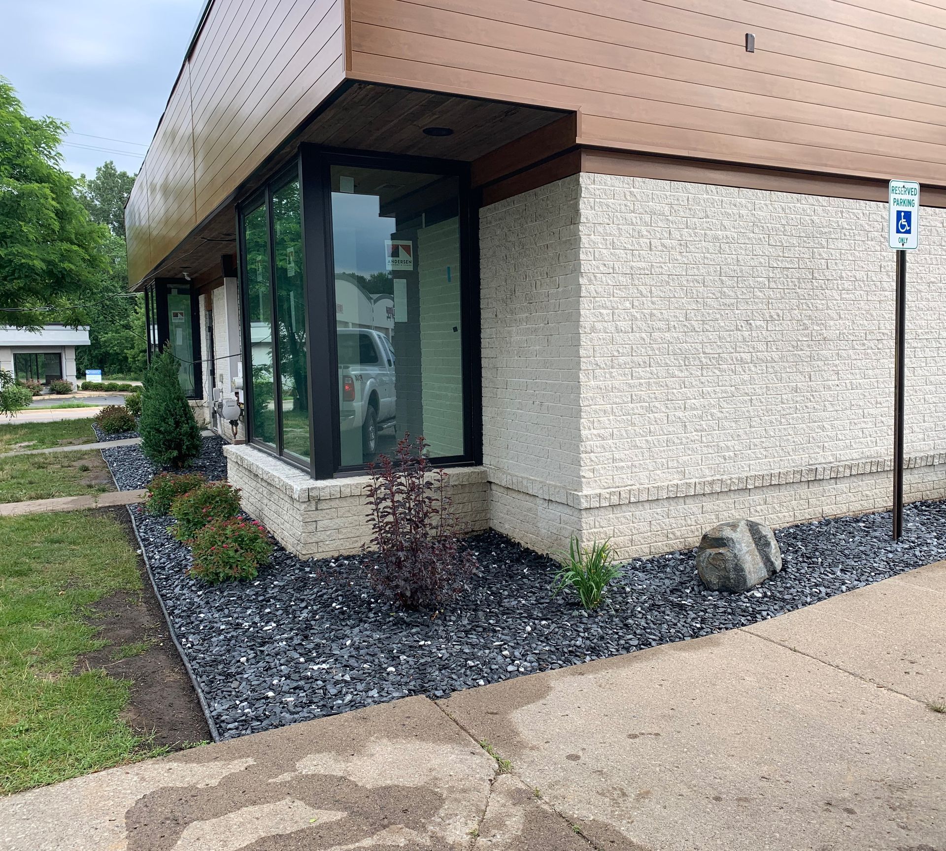Exterior of a building with a wheelchair accessible parking sign. Black mulch bed with bushes, and a large window.