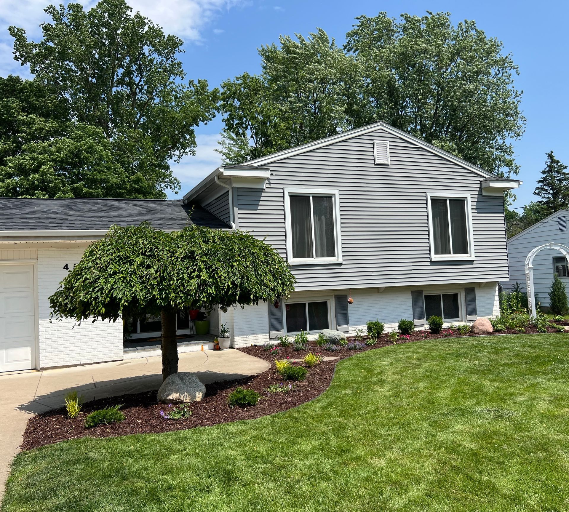 A gray house with a curved bed of mulch and bushes, a weeping tree, and a lawn on a sunny day.
