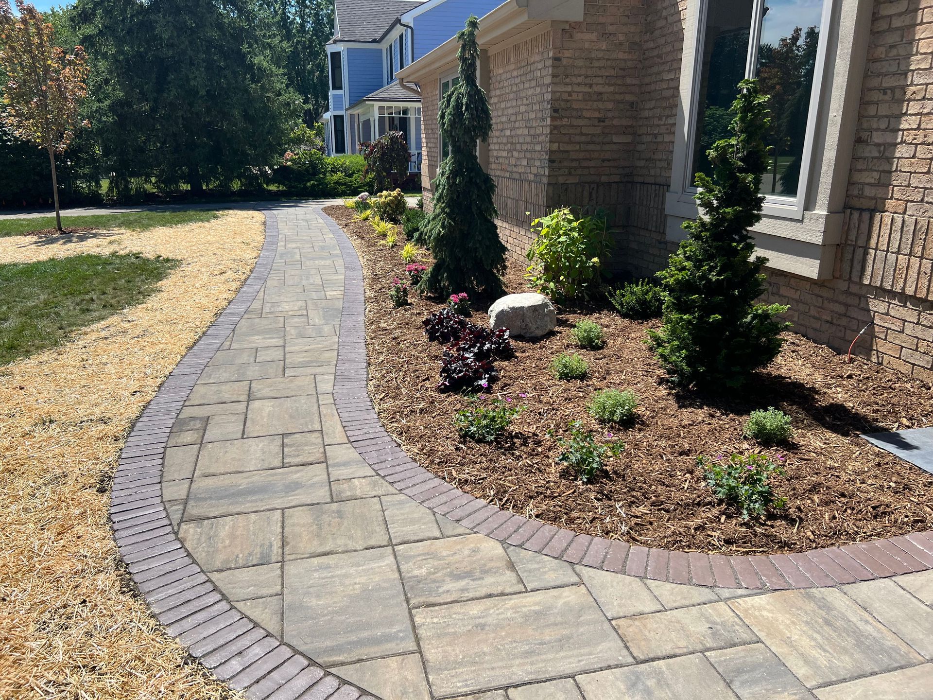 Brick walkway curves past a landscaped flowerbed with brick edging, trees, and a brick home.
