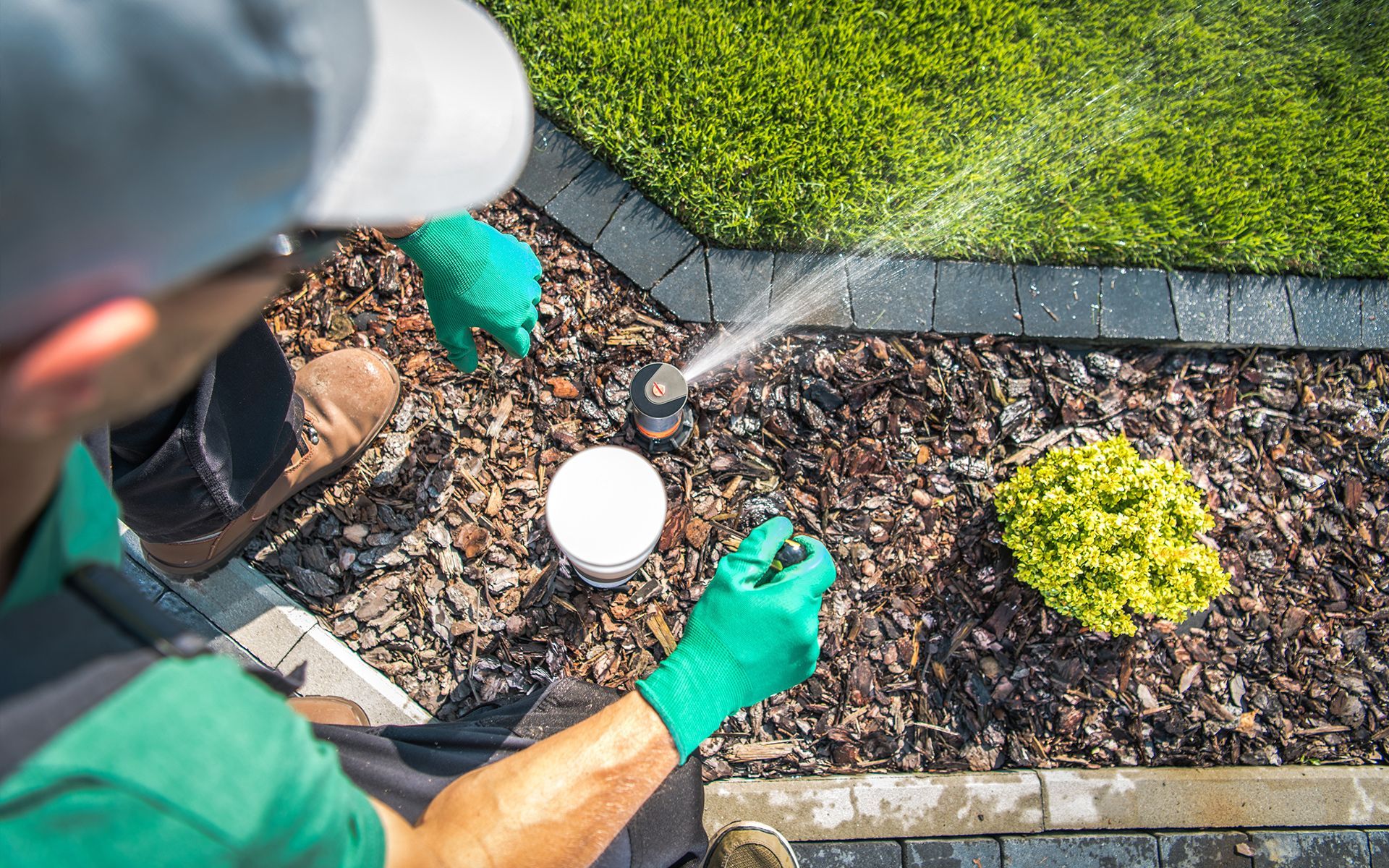 Man in green gloves adjusts sprinkler head in a garden bed with mulch and plants, near grass.