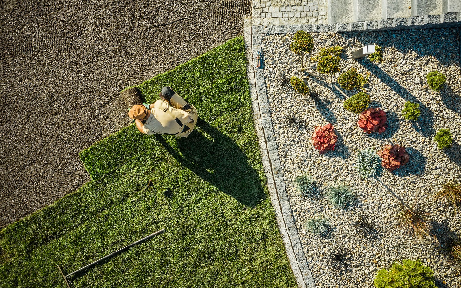 Bird's-eye view of a person in a tan outfit laying sod next to a rock garden with colorful plants.