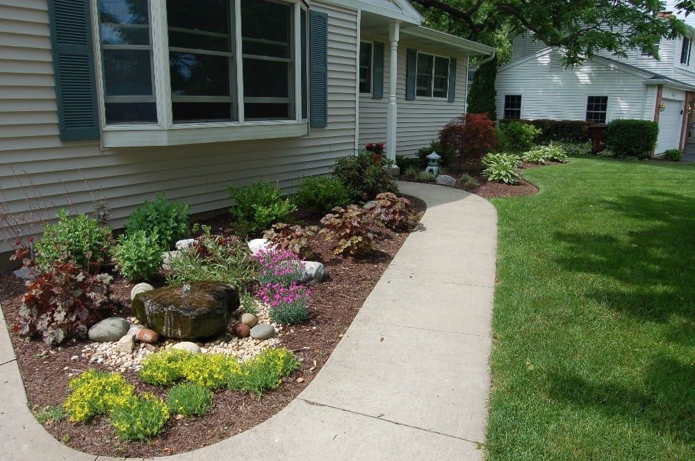 A landscaped front yard with a curving walkway, various plants, and a rock fountain in front of a house.