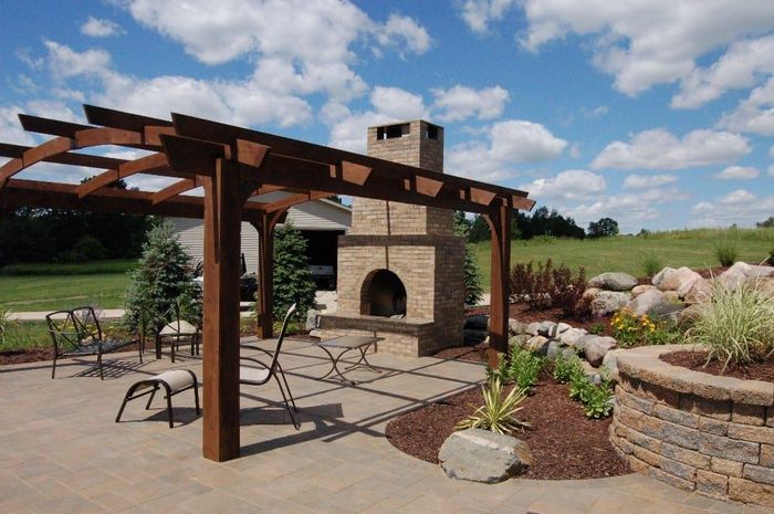Patio with pergola, fireplace, seating, and landscaping under a blue sky with clouds.