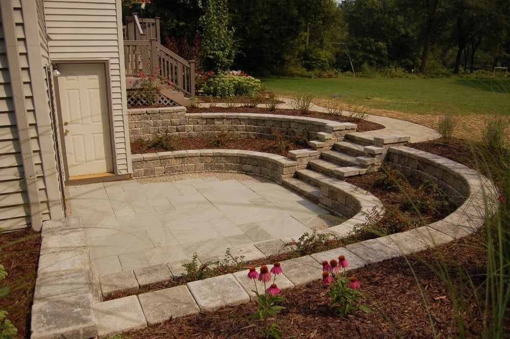 Stone patio with curved retaining walls, steps, and a door on the side of a house.