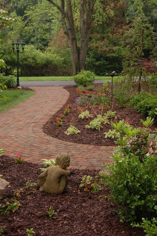Brick pathway winds through a garden with a statue, flowers, and trees.