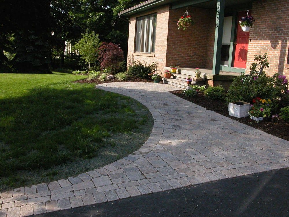 Brick pathway leads to a brick house with a red door, surrounded by grass and flower beds.