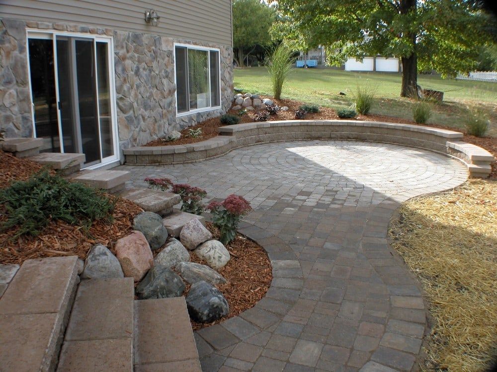 Stone patio with curved edges and retaining wall, next to house with stone veneer and sliding glass door.
