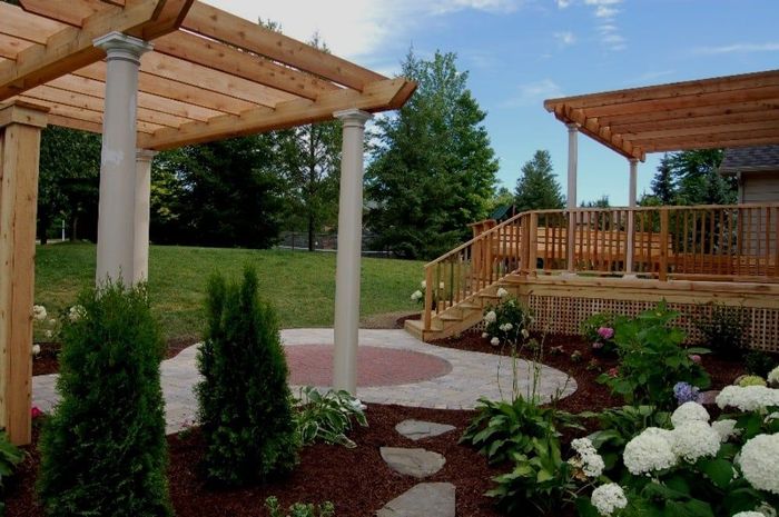 Wooden pergola with stone patio, deck, and garden. Green grass and plants under blue sky.