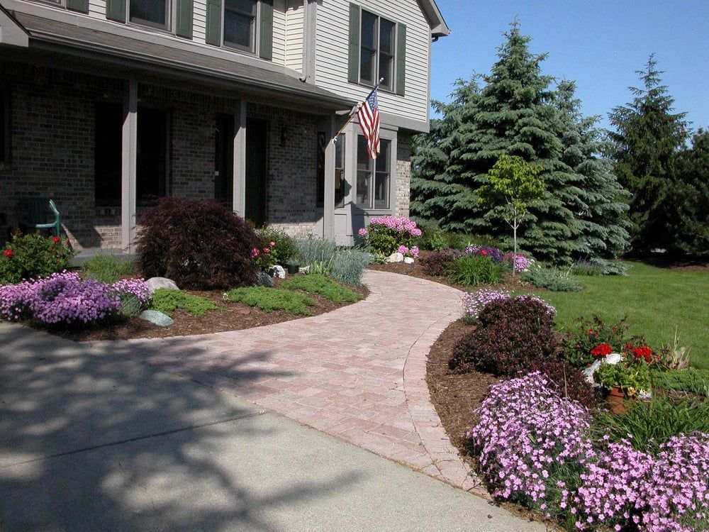 Brick pathway curves toward a house with lush landscaping, including purple flowers and a green lawn.