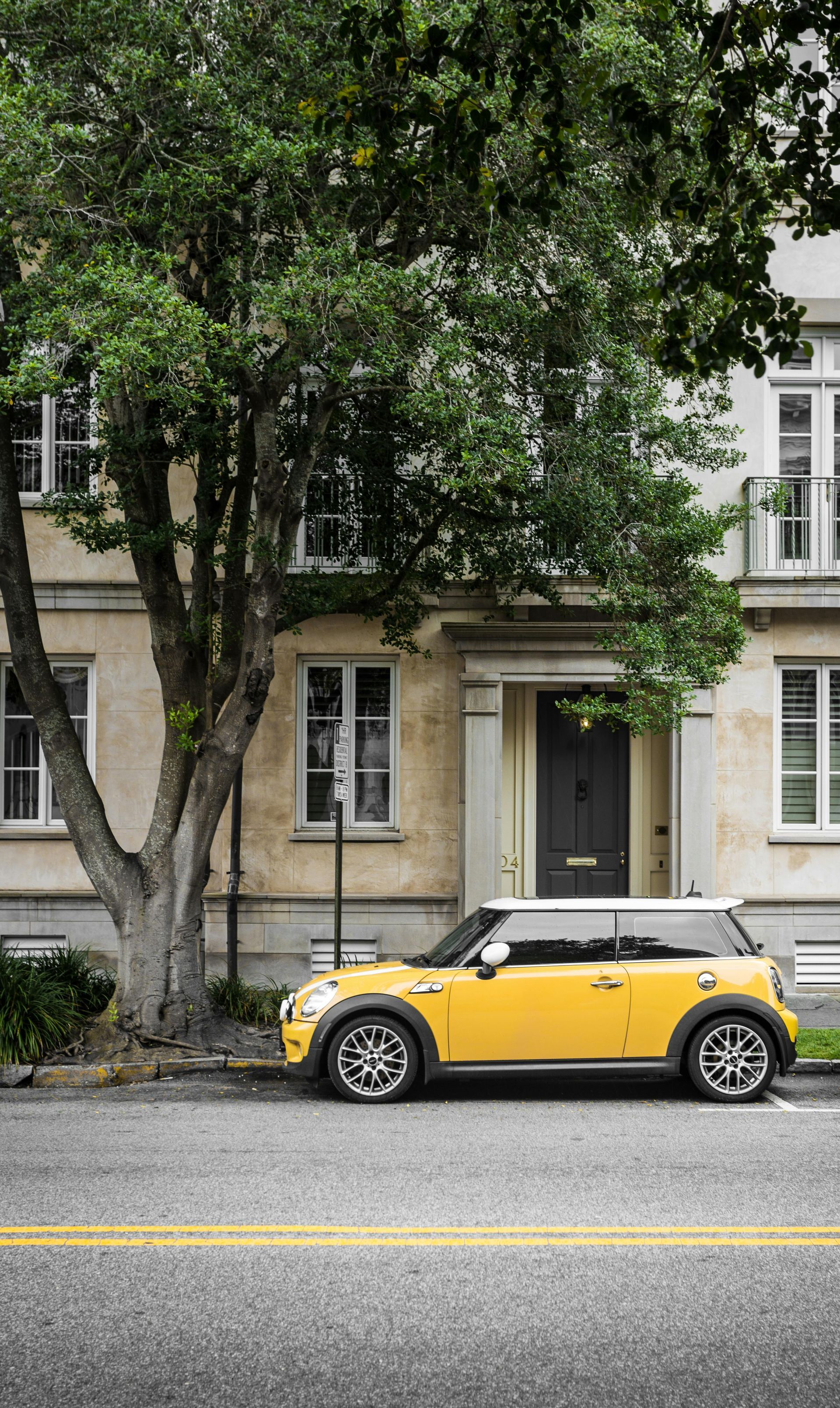 Yellow Mini Cooper parked on a street in front of a beige building with a tree.