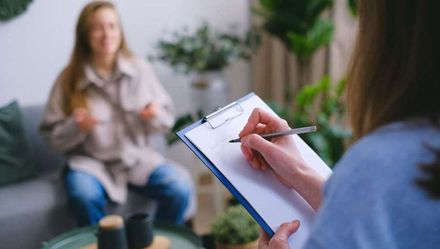 A woman is sitting on a couch while a woman holds a clipboard.